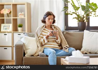 health, cold and people concept - sad sick young woman in blanket sitting on sofa and drinking hot tea at home. sad sick young woman drinking hot tea at home