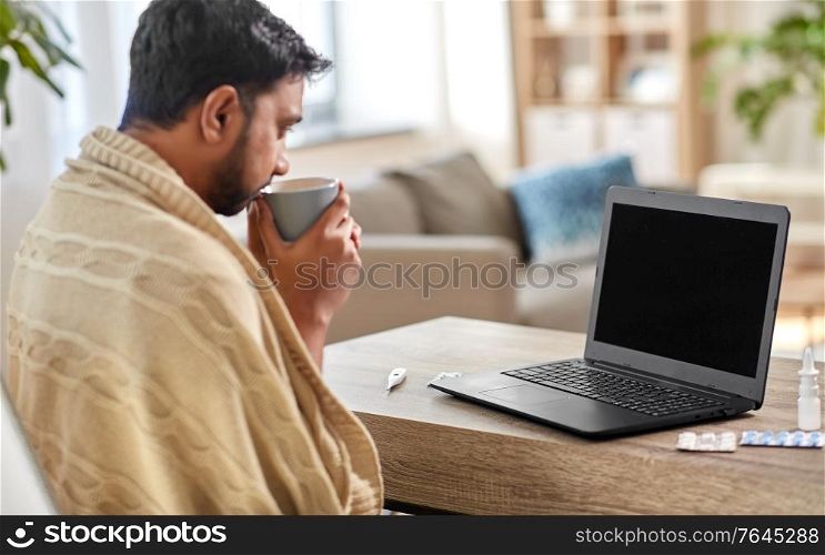 health, cold and people concept - sick young indian man in blanket with laptop computer drinking hot tea and having video call at home. sick indian man with laptop drinking tea at home