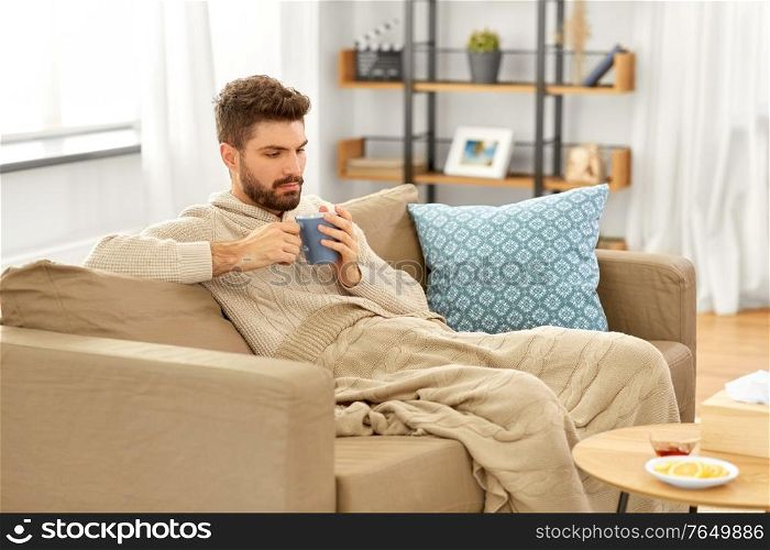 health, cold and people concept - sick young man in blanket drinking hot tea with lemon and honey at home. sick young man in blanket drinking hot tea at home