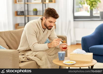 health, cold and people concept - sick young man in blanket drinking hot tea with lemon and honey at home. sick young man in blanket drinking hot tea at home