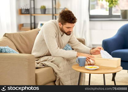 health, cold and people concept - sick young man in blanket taking paper tissue from box with hot tea, lemon and honey on table at home. sick man taking paper tissue from box at home