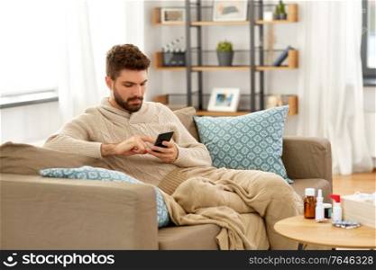 health, cold and people concept - sick young man in blanket with smartphone and medicine at home. sick young man in blanket with smartphone at home