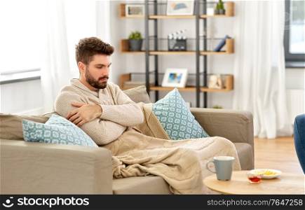 health, cold and people concept - sick young man in blanket with hot tea, lemon and honey on table at home. sick young man in blanket with hot tea at home