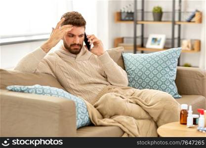 health, cold and people concept - sick young man in blanket with smartphone and medicine calling doctor at home. sick young man calling on smartphone at home