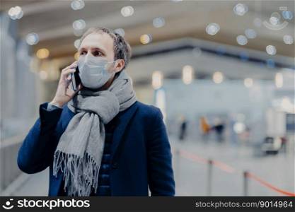 Health, communication and epidemy concept. Serious man wears protective medical mask, cares about safety not to catch coronavirus, walks in shopping mall, talks via cell phone. Anti virus protection