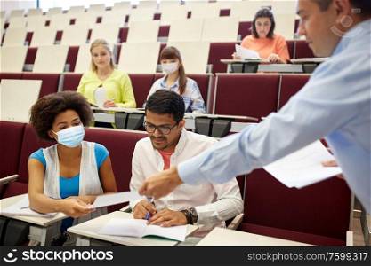 health, education and pandemic concept - teacher giving tests to students wearing protective medical mask for protection from virus at lecture hall. teacher and students in masks at lecture hall