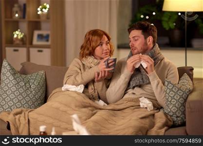 health, flu and people concept - sick young couple in scarves drinking hot tea and blowing nose at home. sick young couple drinking hot tea at home