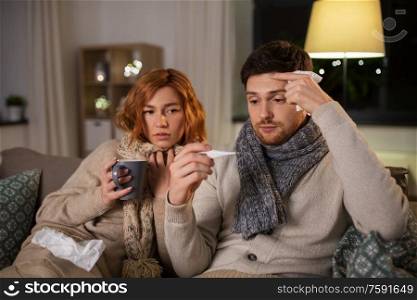 health, flu and people concept - sick young couple in scarves with thermometer measuring temperature and drinking hot tea at home. sick couple with thermometer and tea at home