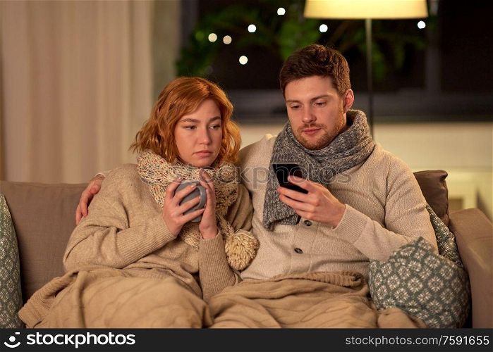 health, flu and people concept - sick young couple in scarves with smartphone drinking tea at home. sick young couple with smartphone at home