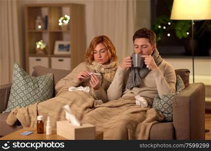 health, flu and people concept - sick young couple in scarves with thermometer measuring temperature and drinking hot tea at home. sick couple with thermometer and tea at home