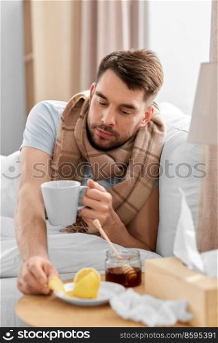 health, illness and people concept - sick man drinking hot tea with lemon and honey in bed at home. sick man drinking hot tea in bed at home
