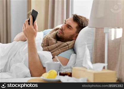 health, illness and people concept - sick man with smartphone in bed at home. sick man with smartphone in bed at home