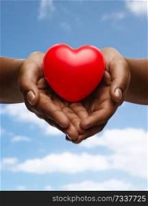 health, medicine and charity concept - close up of african american female hands with small red heart over blue sky and clouds background. female hands with small red heart