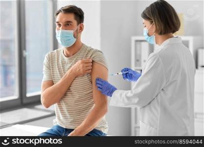 health, medicine and pandemic concept - female doctor or nurse wearing protective medical mask and gloves with syringe vaccinating male patient at hospital. doctor with syringe vaccinating male patient