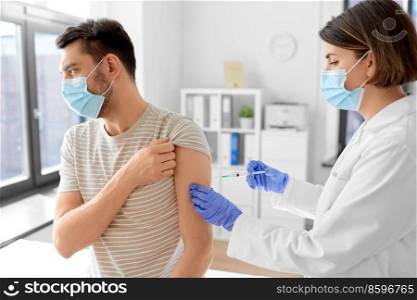 health, medicine and pandemic concept - female doctor or nurse wearing protective medical mask and gloves with syringe vaccinating male patient at hospital. doctor with syringe vaccinating male patient