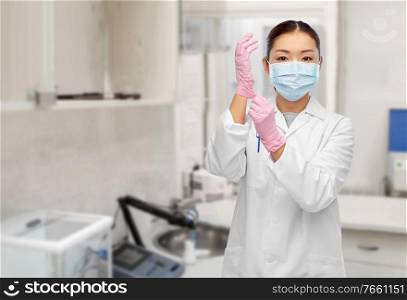 health, medicine and pandemic concept - young asian female doctor or scientist wearing face protective medical mask and gloves for protection from virus disease over hospital or laboratory background. asian female doctor in medical mask and gloves