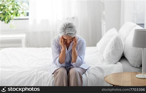 health, old age and people concept - senior woman in pajamas suffering from headache sitting on bed at home bedroom. senior woman with headache sitting on bed at home