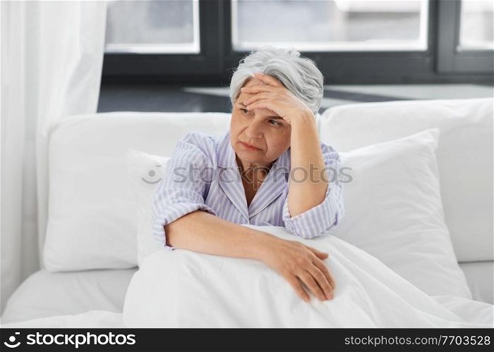 health, old age and people concept - senior woman in pajamas suffering from headache sitting in bed at home bedroom. senior woman with headache sitting in bed at home