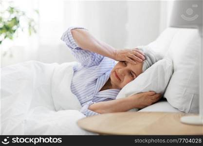 health, old age and people concept - senior woman in pajamas suffering from headache lying in bed at home bedroom. senior woman with headache in bed at home