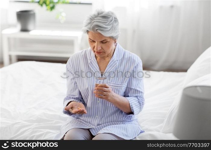 health, old age and people concept - senior woman in pajamas with medicine pill and glass of water sitting on bed at home bedroom. senior woman with pill and water sitting on bed