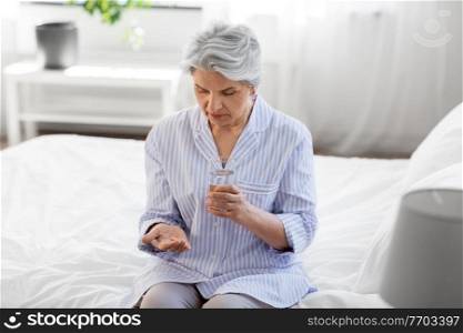 health, old age and people concept - senior woman in pajamas with medicine pill and glass of water sitting on bed at home bedroom. senior woman with pill and water sitting on bed