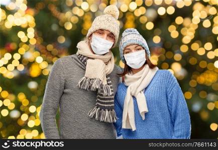health, people and winter holidays concept - couple wearing face protective medical mask for protection from virus disease in knitted hats and scarves over christmas tree lights background. couple in masks and winter clothes on christmas