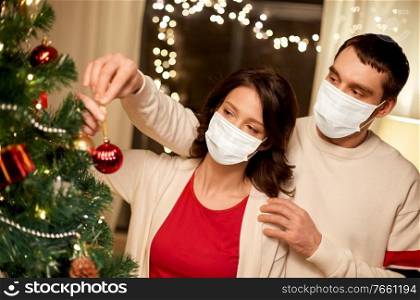 health, people and winter holidays concept - couple wearing face protective medical mask for protection from virus disease decorating christmas tree at home. couple in masks decorating christmas tree at home