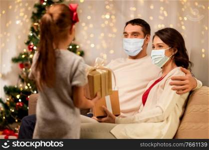 health, people and winter holidays concept - mother, father wearing face protective medical mask for protection from virus disease and little daughter with christmas present at home. family in masks with christmas presents at home