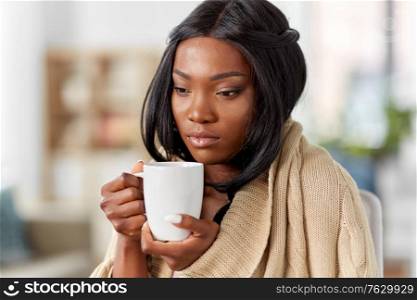health problem and people concept - young sick african amercian woman in blanket drinking hot tea at home. sick african amercian woman drinking tea at home