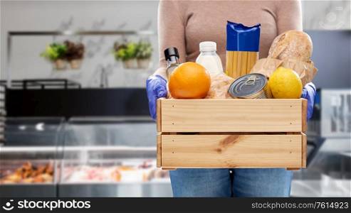 health protection, safety and pandemic concept - close up of woman in gloves with food in wooden box over grocery store background. woman in gloves with food in wooden box