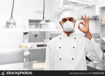 health protection, safety and pandemic concept - indian male chef cook wearing face protective mask or respirator showing ok gesture over restaurant kitchen background. chef in respirator showing ok sign at kitchen