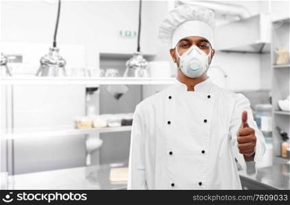 health protection, safety and pandemic concept - indian male chef cook wearing face protective mask or respirator showing thumbs up over restaurant kitchen background. chef in respirator showing thumbs up at kitchen