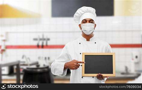 health protection, safety and pandemic concept - indian male chef cook wearing face protective mask with blank chalkboard over restaurant kitchen background. chef in face mask with chalkboard at kitchen
