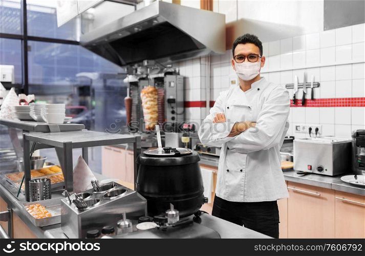 health protection, safety and pandemic concept - male chef cook wearing face protective medical mask over kebab shop kitchen background. male chef with in face mask at kebab shop kitchen