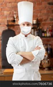 health protection, safety and pandemic concept - male chef cook with crossed hands wearing face protective medical mask for protection from virus disease over restaurant kitchen background. male chef in face mask at restaurant kitchen