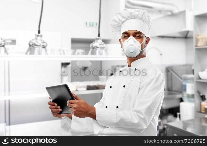 health protection, safety and pandemic concept - male indian chef in toque wearing face protective mask or respirator with tablet computer over restaurant kitchen background. chef in respirator with tablet pc at kitchen