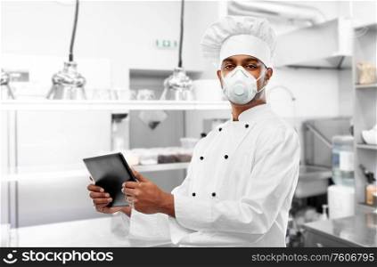 health protection, safety and pandemic concept - male indian chef in toque wearing face protective mask or respirator with tablet computer over restaurant kitchen background. chef in respirator with tablet pc at kitchen