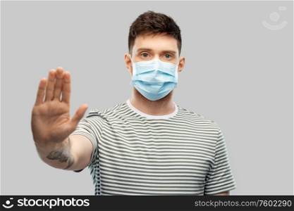 health protection, safety and pandemic concept - young man in protective medical mask making stop gesture over grey background. man in protective medical mask making stop gesture