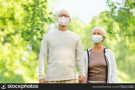 health, quarantine and pandemic concept - senior couple wearing protective medical mask for protection from virus holding hands outdoors over green natural background. senior couple in protective medical masks outdoors