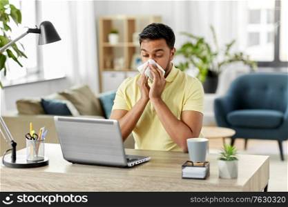 health, remote job and people concept - sick young indian man with laptop computer blowing nose at home office. indian man with laptop blowing nose at home office