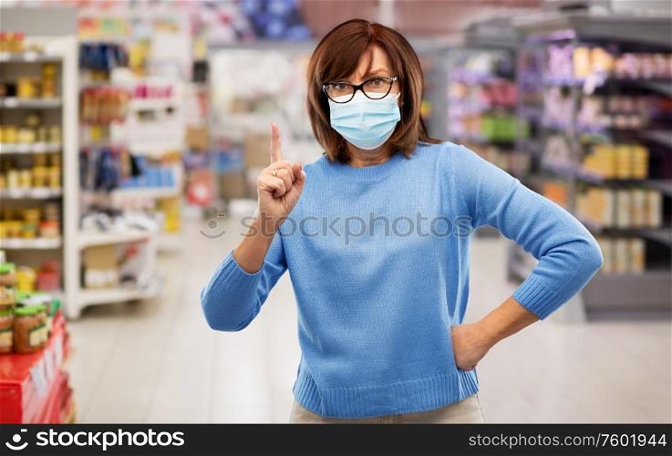 health, safety and pandemic concept - angry senior woman in glasses wearing protective medical mask for protection from virus and pointing finger up over supermarket background. angry senior woman in medical mask at supermarket