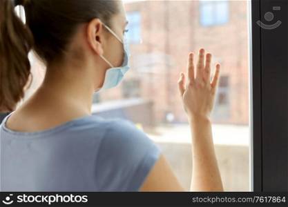 health, safety and pandemic concept - close up of sick young woman wearing protective medical mask looking through window at home. sick young woman wearing protective medical mask