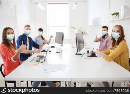 health, safety and pandemic concept - creative team wearing protective medical masks for protection from virus disease with computers showing thumbs up at office. business team in medical masks showing thumbs up