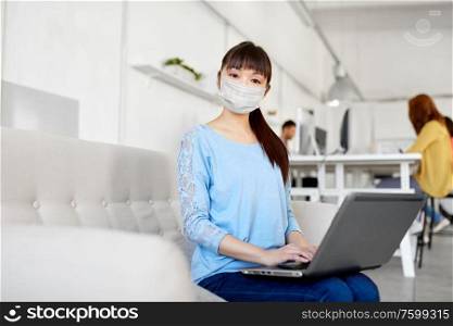 health, safety and pandemic concept - happy young asian woman with laptop computer in protective medical mask for protection from virus and working at office. asian woman with laptop in medical mask at office