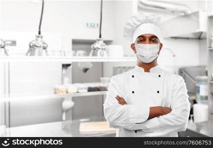 health, safety and pandemic concept - indian male chef cook in toque with crossed hands wearing face protective medical mask for protection from virus disease at restaurant kitchen. male chef in face mask at restaurant kitchen