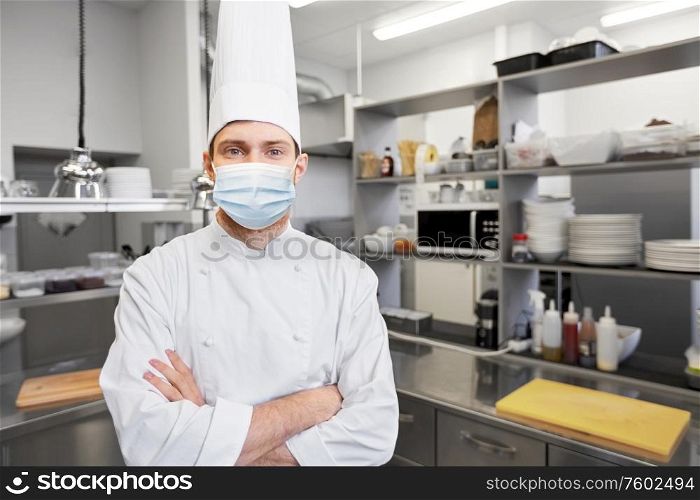 health, safety and pandemic concept - male chef cook in toque with crossed hands wearing face protective medical mask for protection from virus disease at restaurant kitchen. male chef cook in face mask at restaurant kitchen