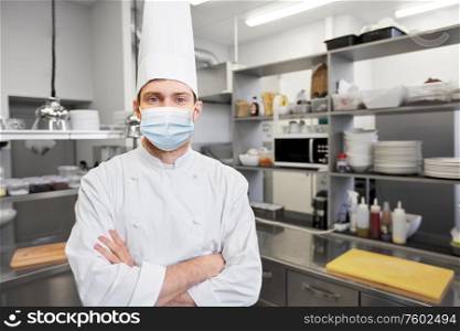 health, safety and pandemic concept - male chef cook in toque with crossed hands wearing face protective medical mask for protection from virus disease at restaurant kitchen. male chef cook in face mask at restaurant kitchen