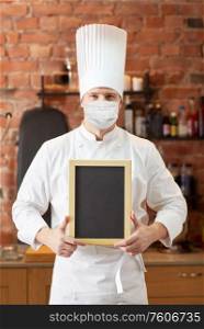 health, safety and pandemic concept - male cook or baker wearing face protective medical mask for protection from virus disease with blank menu chalk board over restaurant kitchen background. male chef in face mask with black menu chalk board