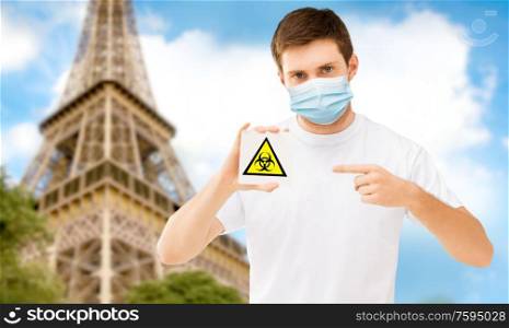 health, safety and pandemic concept - man wearing protective medical mask holding biohazard caution sign over eiffel tower in paris, france background. man in mask with biohazard sign in france