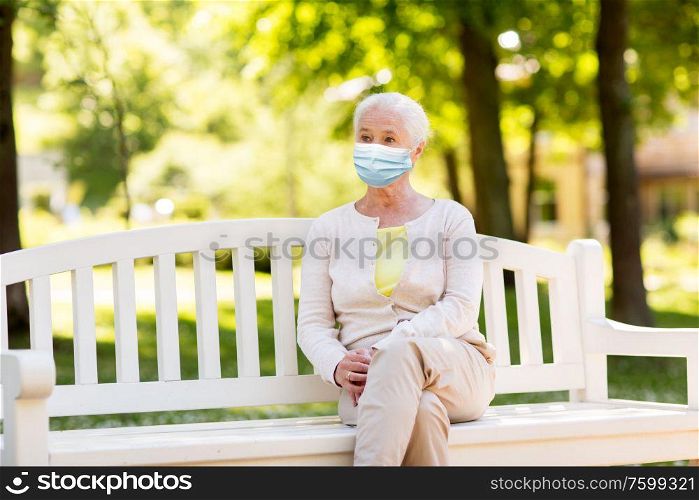 health, safety and pandemic concept - sad senior woman wearing protective medical mask for protection from virus sitting on bench at summer park. senior woman in protective medical mask at park